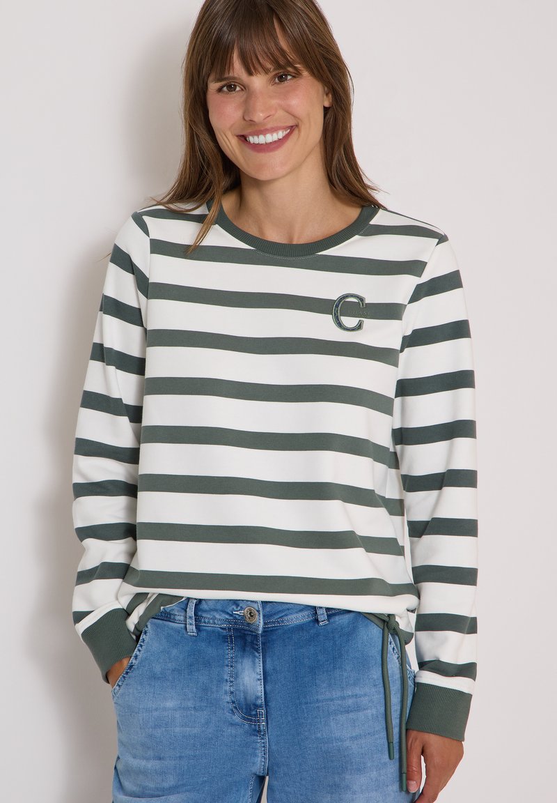 Femme souriante portant une chemise à manches longues rayée verte et blanche avec un emblème "C" et un jean bleu, les mains dans les poches.
