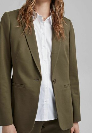Vrouw draagt een olijfgroene blazer over een wit overhemd met knopen, getoond van nek tot heup tegen een lichtgrijze achtergrond.