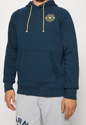 Mann trägt eine marineblaue Kapuzenjacke mit gelben Kordeln und dem "Seven Bucks"-Logo auf der Brust, kombiniert mit hellgrauen Jogginghosen.