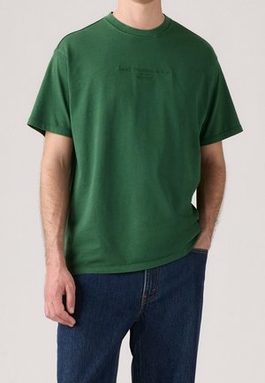 Homme portant un T-shirt Levi's vert foncé à manches courtes avec un jean bleu foncé, debout avec une main dans la poche et l'autre détendue.