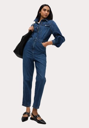 Combinaison en denim avec col, fermeture éclair et manches bouffantes. Portée avec des chaussures à talons noires avec une lanière arrière et un sac fourre-tout noir texturé.