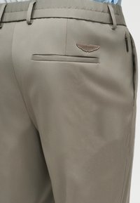 Κοντινό πλάνο στην τσέπη μπεζ παντελονιού με θηλιές για ζώνη και ραμμένη πάνω από την πίσω τσέπη ετικέτα με το λογότυπο της Aston Martin.