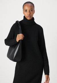 Črna rebrasta pulover majica s turtleneck ovratnikom in dolgimi rokavi, v kombinaciji z gladko črno usnjeno torbico za ramo, ki jo drži ena roka pred enotno ozadjem.