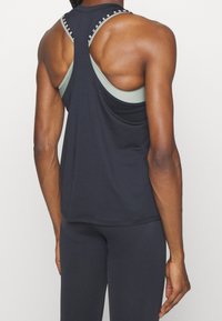 Top deportivo negro sin mangas con diseño de espalda de nadador, que presenta una capa inferior de color verde claro y acentos de marca a lo largo del escote.