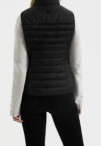 Svart puffer-väst med horisontell quiltad design, hög krage och tryckknappsstängning. Bärs över en ljusgrå tröja och svarta jeans.