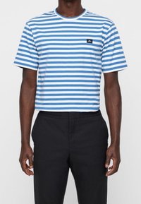 Muž v modro-bíle pruhovaném tričku s krátkým rukávem a černých kalhotách, stojící s uvolněnýma rukama podél těla.