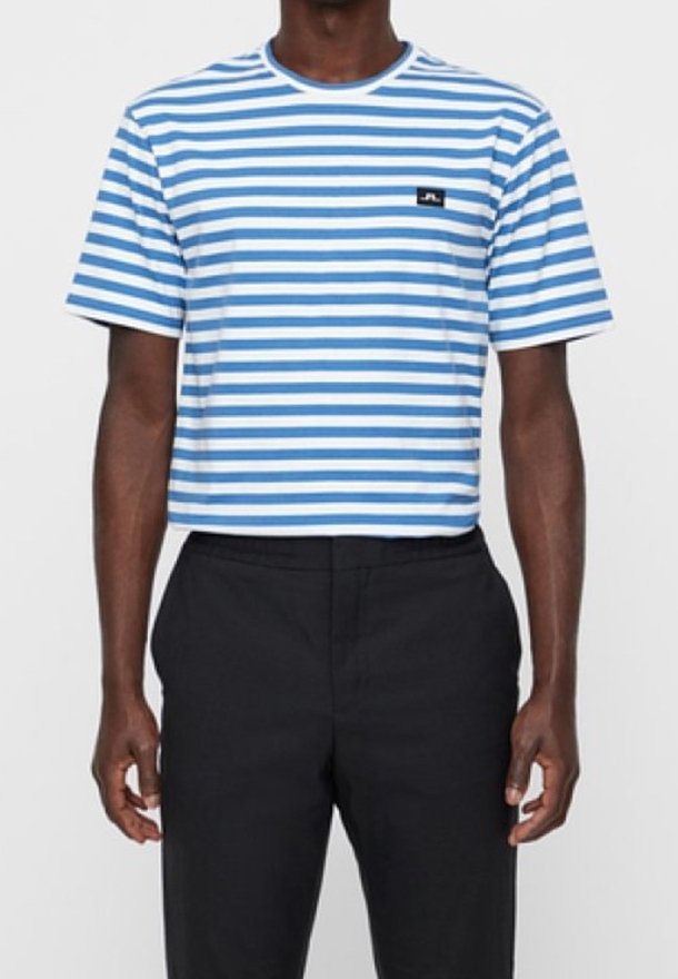 Muž v modro-bíle pruhovaném tričku s krátkým rukávem a černých kalhotách, stojící s uvolněnýma rukama podél těla.