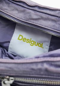 Fioletowe wnętrze torby z materiału, z metalowym suwakem. Zawiera białą metkę z zielonym napisem "Desigual" oraz gładką strukturę.
