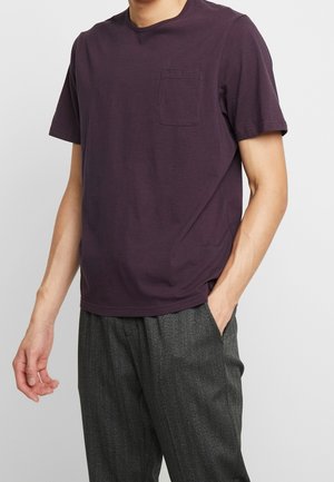 Kurzärmeliges T-Shirt mit Rundhalsausschnitt aus tiefpurpurfarbener Baumwolle mit Brusttasche; kombiniert mit grauen strukturierten Hosen.