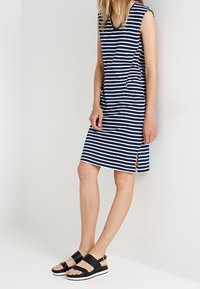 Kvinna klädd i en ärmlös marinblå och vit randig knälång klänning med sidoklipp och svarta platåsandaler, stående mot en vit vägg.