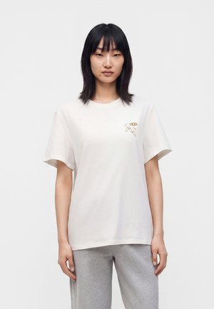 Mujer con cabello negro liso que lleva una camiseta blanca de manga corta UGG y pantalones gris claro, de pie frente a un fondo blanco liso.