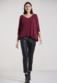 Burgunderfarbener V-Ausschnitt-Pullover mit 3/4-Ärmeln, kombiniert mit schwarzen Kunstlederhosen und Ankle Boots, vor einem neutralen Hintergrund präsentiert.