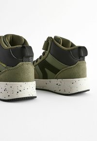 High-top sneakers met een olijfgroen en zwarte kleurenschema, gemaakt van suède en mesh materialen, met een gespikkelde witte zool en een verstelbare band.