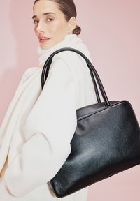 Borsa a tracolla in pelle nera con texture liscia, forma rettangolare e due manici robusti, posizionata su uno sfondo rosa chiaro.