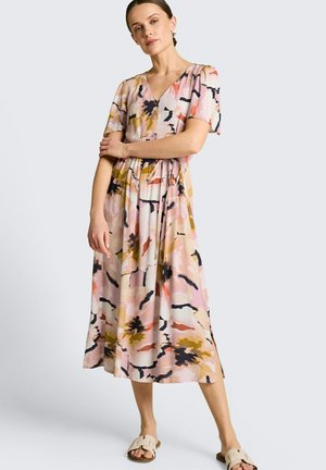 Frau trägt ein Midi-Kleid mit abstraktem Muster in Pink, Senf und Schwarz, kurzen Ärmeln und beigen Slip-On-Sandalen, steht mit verschränkten Armen.