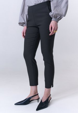 Person, die eng anliegende schwarze, knöchellange Hose, eine graue Bluse mit Puffärmeln und schwarze spitze Slingback-Absätze trägt, steht auf einem hellen Boden.