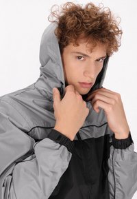 Giacca leggera con cappuccio grigia e nera. Presenta una zip frontale, polsini elasticizzati e due cordini regolabili sul cappuccio.