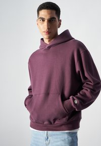 Sudadera con capucha color marrón, con un bolsillo tipo canguro y acentos en los puños. La tela parece suave, con un corte holgado y un diseño minimalista.
