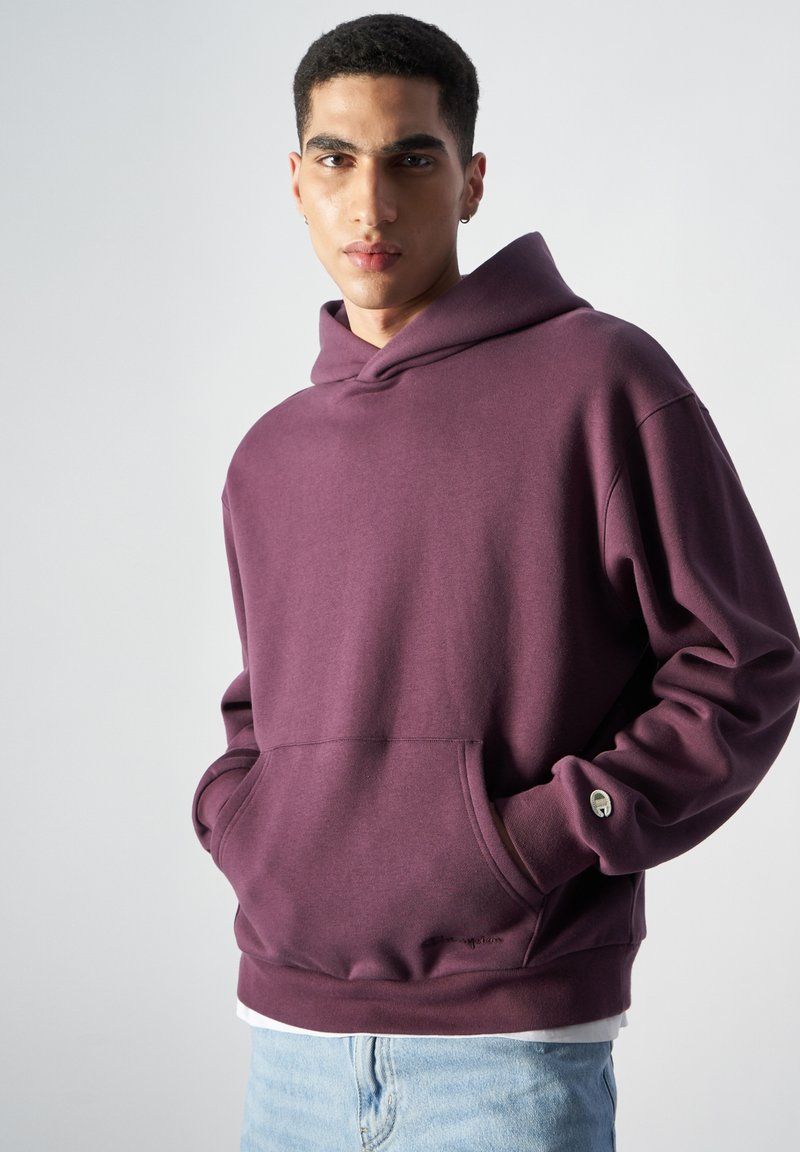 Sudadera con capucha color marrón, con un bolsillo tipo canguro y acentos en los puños. La tela parece suave, con un corte holgado y un diseño minimalista.