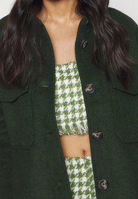 Green wool blend jacket with large buttons and two chest pockets, worn over a green and white houndstooth cropped top with frayed edges.