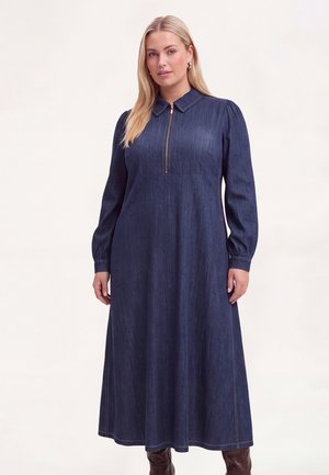 Frau trägt ein langes dunkelblaues Jeanskleid mit Frontreißverschluss und Kragen, kombiniert mit braunen Stiefeln, steht vor einem schlichten Hintergrund.