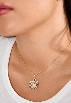 Close-up van een persoon die een gouden spiraalvormige hanger draagt aan een dun gouden kettinkje, met een wit topje zichtbaar bij het sleutelbeen.