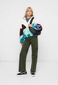 Pullover in pile colorato con colletto alto, abbinato a pantaloni larghi verde oliva e sneakers nere, porta una borsa a spalla nera.
