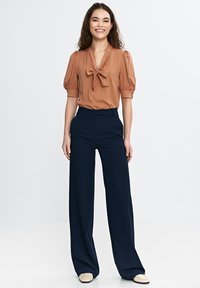 Brązowa bluzka z kokardą, krótkie bufiaste rękawy. Granatowe spodnie o szerokich nogawkach, z zaznaczonym przodem. Beżowe buty wsuwane. Prosty, dopasowany fason.
