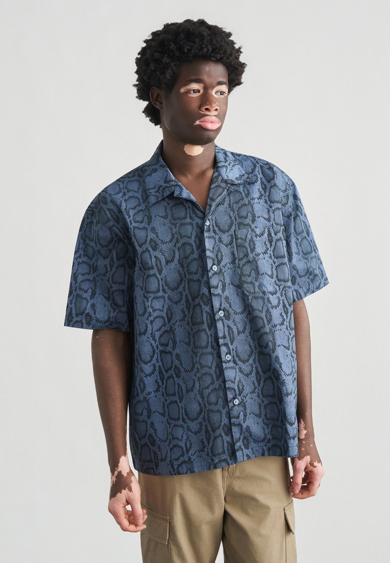 Camisa de manga corta con botones, presenta un patrón de piel de serpiente en azul oscuro, fabricada con tela texturizada. Corte casual con cuello abierto y bolsillo.