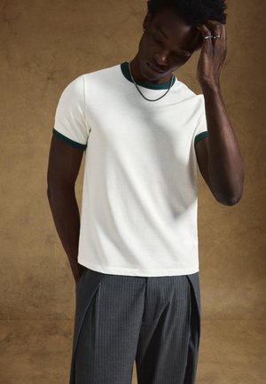 Witte T-shirt met korte mouwen en donkergroene bies, gecombineerd met donkere pantalon met fijne strepen. Gladde stof, comfortabele pasvorm, zonder zichtbare patronen.