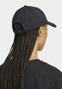 Czarny bawełniany kapelusz z zakrzywionym daszkiem, regulowanym paskiem z tyłu i widocznymi detalami w szyciu. Noszony z długimi zaplecionymi włosami.