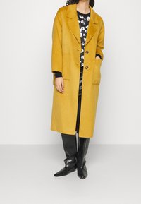 Žena ve dlouhém hořčicově žlutém kabátě přes černý květovaný top, černou koženou sukni a černé kozačky sahající ke kolenům, stojící před jednobarevným pozadím.