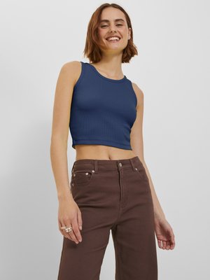 Smilende ung kvinne iført en ermeløs marineblå cropped topp og høytlivs brune bukser, stående foran en ensfarget lys bakgrunn.