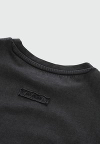 Schwarzes Baumwoll-T-Shirt mit Rundhalsausschnitt und einem strukturierten Patch-Label, das auf den Rücken genäht ist. Weicher Stoff mit glatter Oberfläche.