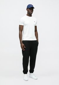 Homme portant un T-shirt blanc à manches courtes, un pantalon de survêtement noir, des baskets blanches et une casquette de baseball bleu marine, debout devant un fond uni.