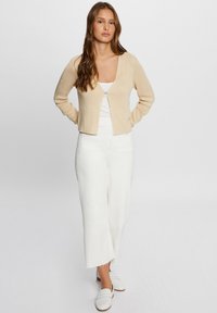 Cardigan beige à côtes, ouvert devant avec un bouton, porté avec un haut blanc et un pantalon à jambes larges. Texture lisse et détails de conception minimaux.