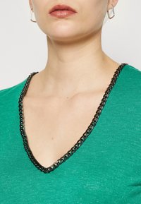 Camisa verde con escote en V y un detalle de cadena negra a lo largo del cuello, hecha de una tela suave con una sutil textura brillante.