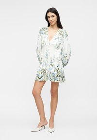 Femeie purtând o rochie mini albă cu flori, mâneci lungi bufante și pantofi cu toc ascuțit albi, stând în fața unui fundal simplu.