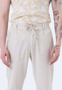 Pantalons en lin beige clair avec une taille à cordon, des poches zippées latérales et une coupe décontractée. Texture lisse et design simple.