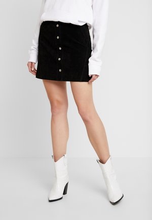 Mujer con un top blanco de manga larga, minifalda negra con botones en la parte delantera y botas blancas de tacón hasta la mitad de la pantorrilla, de pie frente a un fondo liso.