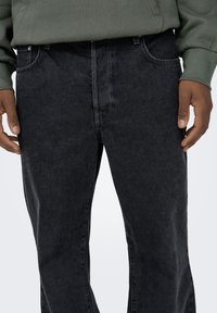 Black denim jeans with a straight leg design, visible front pockets, and contrasting stitching details, shown being worn with a green sweatshirt.
