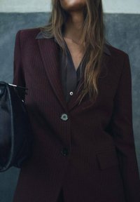 Burgundský blazer s jemným proužkem, s vroubkovaným límcem a stříbrnými knoflíky, nošený přes tmavou košili, držící se černou kabelkou na jedné straně.
