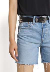 Persona que lleva pantalones cortos de mezclilla azul claro con bordes deshilachados, un cinturón negro con hebilla decorativa plateada y una camiseta negra.