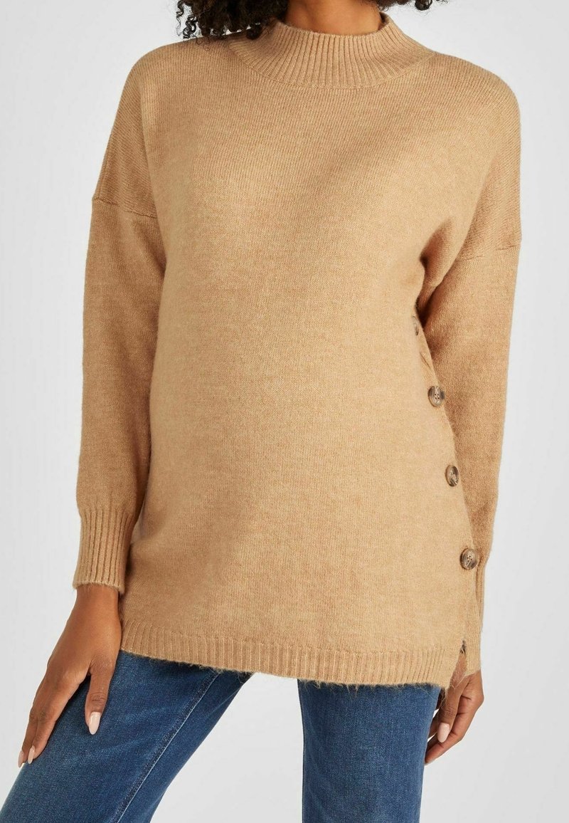Pull en maille marron avec détails de boutons sur le côté porté avec un jean bleu, montré sur une personne du cou jusqu'au milieu de la cuisse.