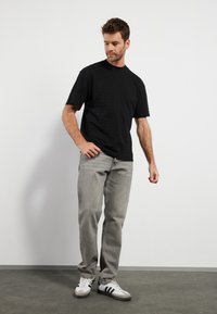 Svart-stripete kortermet t-skjorte, grå jeans og hvite joggesko med svarte detaljer. Enfarget bakgrunn, modell stående med en hånd på hoften.