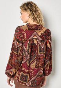Langärmlige Bluse mit mehrfarbigem Design in Rot, Orange und Schwarz, mit komplexen geometrischen Formen und floralen Akzenten. Strukturierter Stoff.