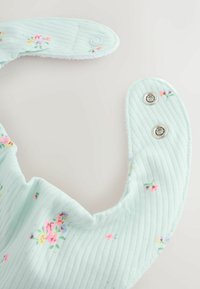 Pehmeä mintunvihreä vauvanruokalappu, jossa on pieni kukkakuvio ja painonapilliset sulkimet vaalealla neutraalilla taustalla.