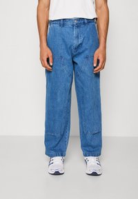 Blue denim cargo pants with loose fit, front pockets, and visible stitching details. Worn with white sneakers and a plain t-shirt.