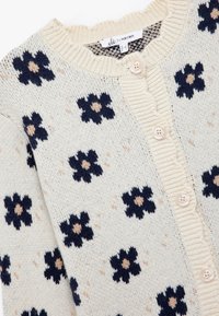 Cárdigan de punto en color crema con un patrón de flores en azul marino y centros en color beige. Presenta un cuello acanalado y un cierre de botones con botones de color claro.