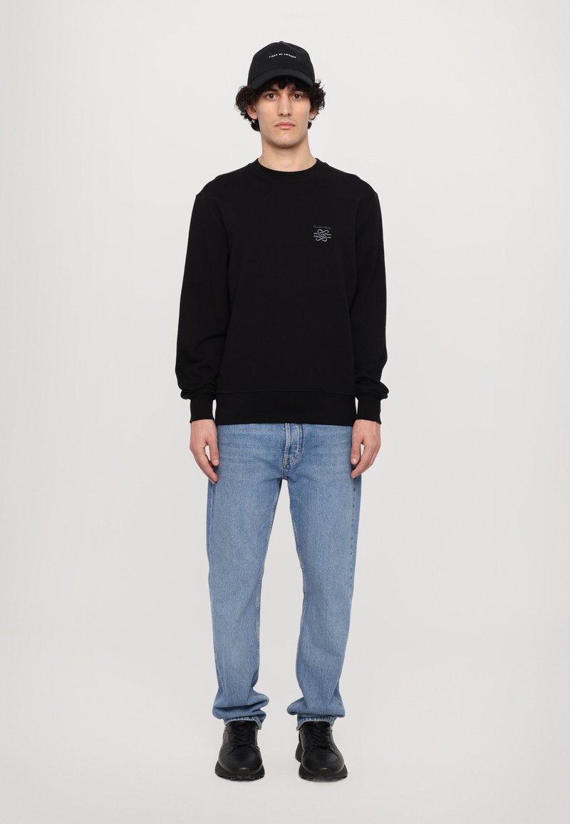 Sweatshirt noir avec un petit motif sur la poitrine, associé à un jean bleu clair et des baskets noires. Le modèle porte une casquette noire.